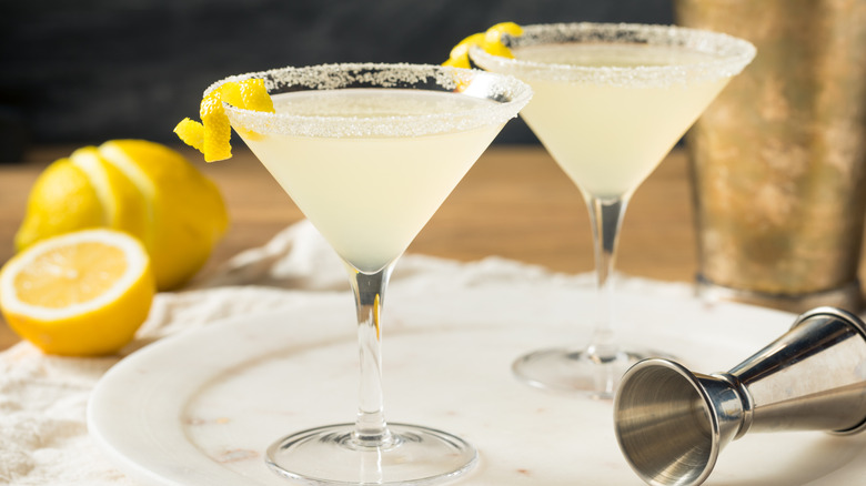 two lemon drop cocktails in martini glasses on a plate next to cut lemons and cocktail measurer