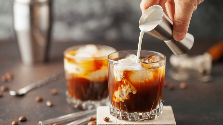 hand pouring cream into White Russian cocktail, second cocktail and cocktail shaker in background