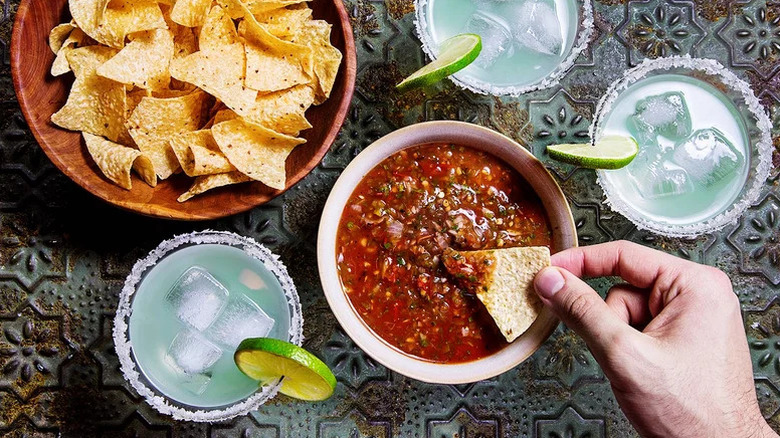 Hand dipping chip into charred tomato salsa with chips and drinks