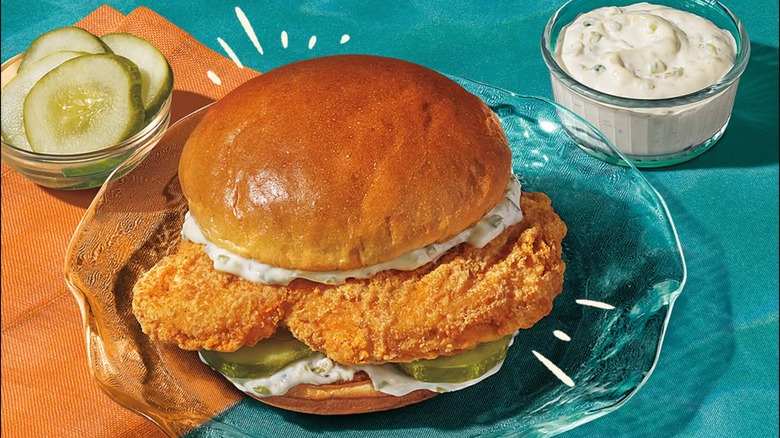 Popeyes flounder fish sandwich on glass plate with pickles and tartar sauce in bowls