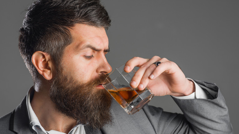 A close up of a person bringing a glass of whiskey to their mouth on a plain background