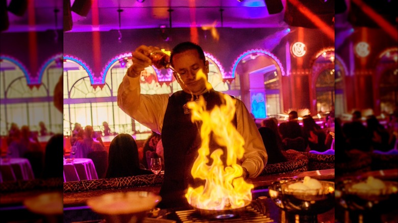 Chef pouring liquid into flaming pan on stove in open kitchen