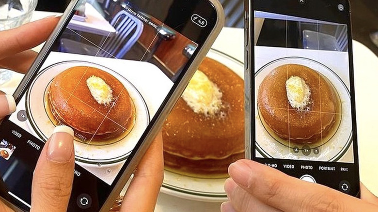 hands holding cellphones taking photos of a stack of pancakes