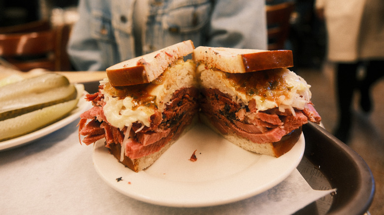 a pastrami sandwich on a white plate with a plate of pickles in the background