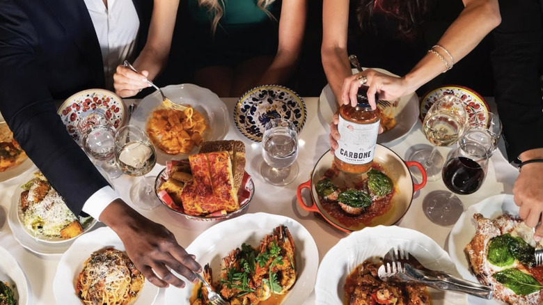a table covered in plates with pasta and other dishes, with hands holding cutlery and picking at the food
