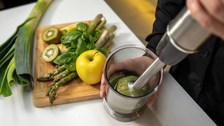 A hand using an immersion blender to blend green ingredients