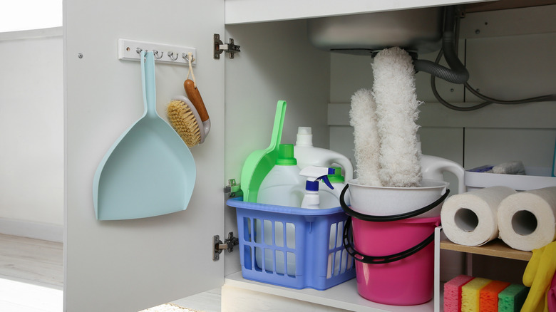 Baskets and bucket with different cleaning supplies and paper towels under sink in kitchen