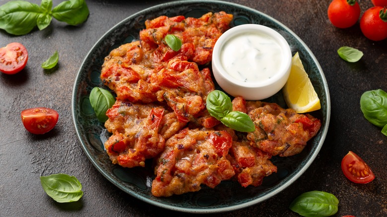 Tomatokeftedes on plate with dip and lemon wedge surrounded by tomatoes and basil