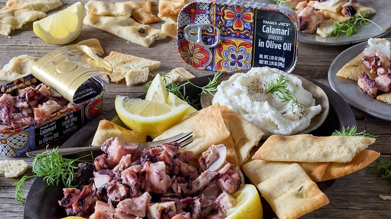 Tins of Trader Joe's Calamari Pieces in Olive Oil on a table surrounded by crackers