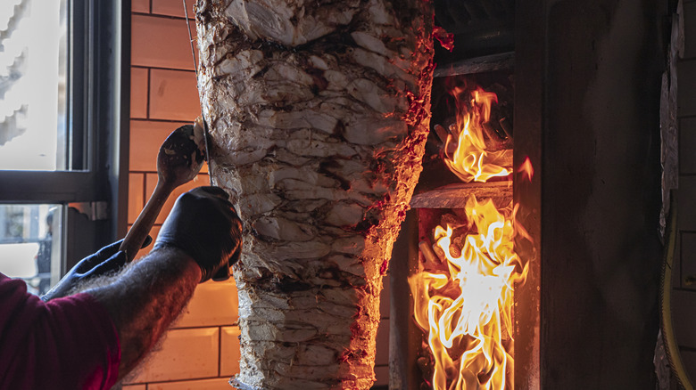 Person slicing döner﻿ kebab meat from vertical rotisserie cooking next to open fire