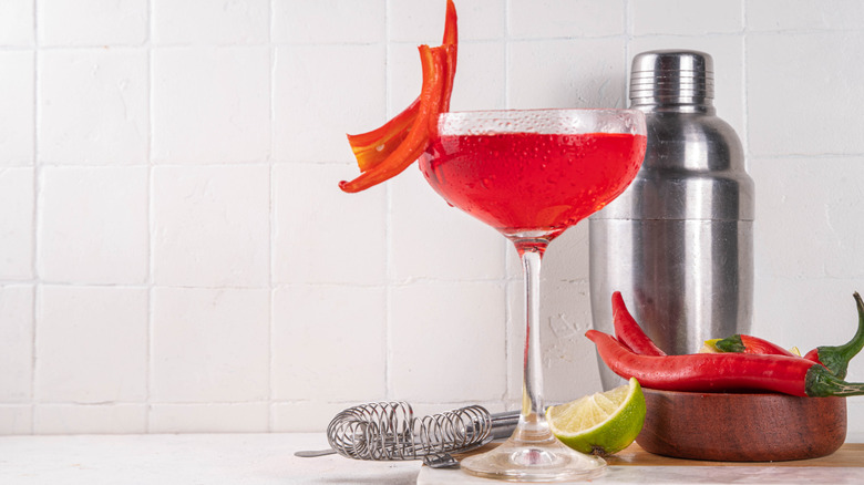 Cocktail served in a coupe glass with chili peppers, lime, and shaker against a white background