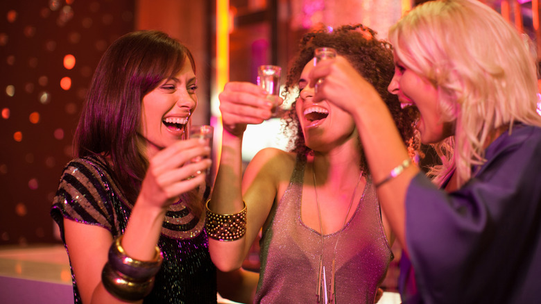 Three friends holding shot glasses and smiling with lights blurred in background