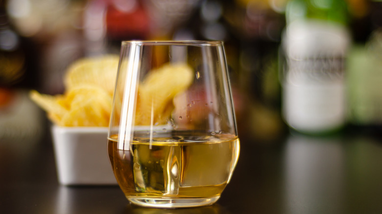 Glass of yellow spirit with snacks and bottles blurred in background