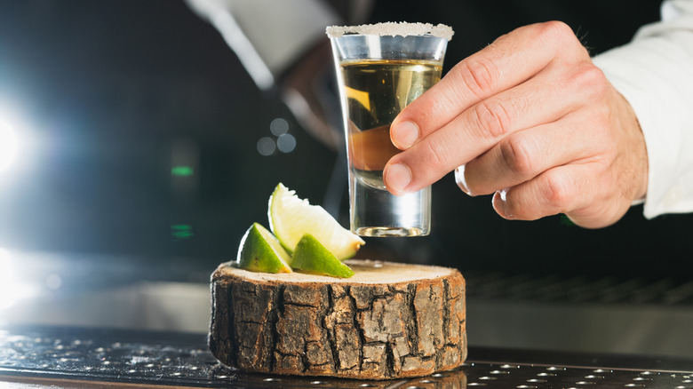 Hand holding shot of tequila over wooden board with slices of limes