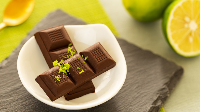 A small bowl of chocolate pieces with lime zest on top. Cut limes are in the background.