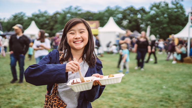 A girl smiles while sticking a fork in a take-out box of food at an outdoor festival