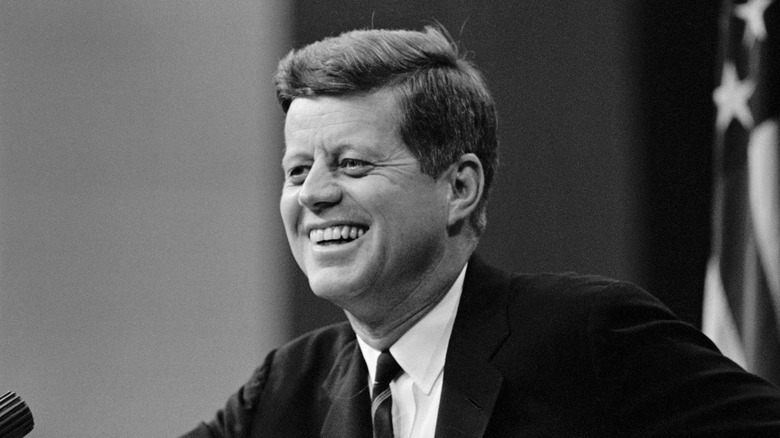 black and white photo of John F. Kennedy smiling with flag in background