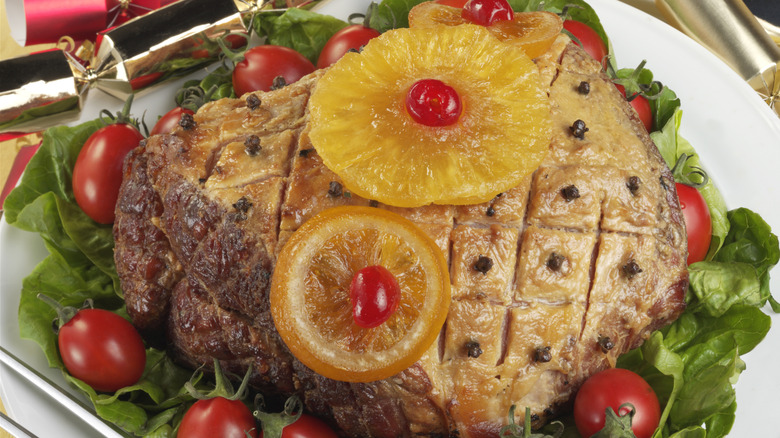 A baked ham with pineapple, cherries, and cloves on a bed of lettuce and grape tomatoes