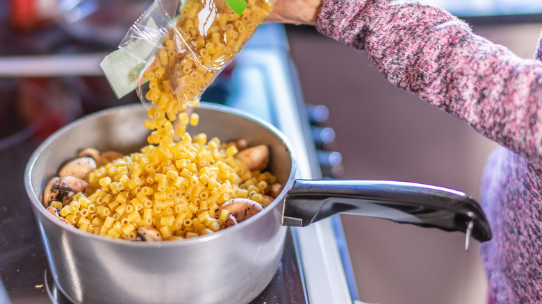 A person pouring a bag of pasta into a pot of food on the stove