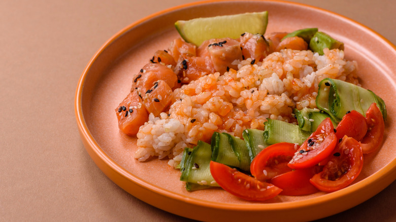 A pink plate of rice served with sliced cucumber, tomatoes, and chunks of salmon