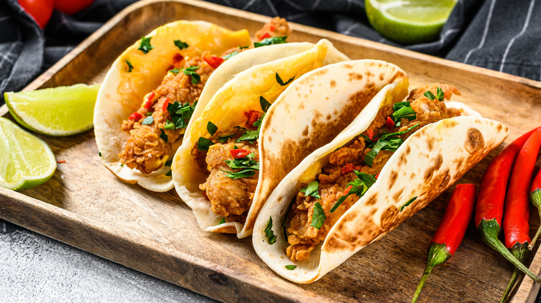 Crispy chicken tacos on wooden serving board with fresh lime wedges and red chili peppers