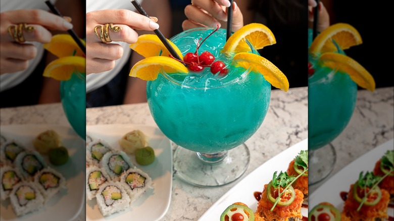 Two hands reaching for two straws in a blue tinted cocktail served in a large glass, garnished with orange wedges and cherries. It sits on a granite table with two plates of sushi.
