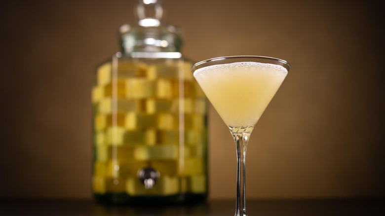 A cloudy yellow liquid in a martini glass. In the background, a glass pitcher with a yellow checker pattern and a metallic spout.