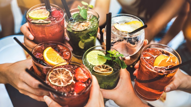 A group of seven people toasting multicolored cocktails garnished with various fruit.