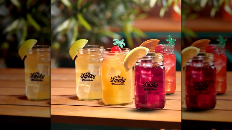 A group of multicolored drinks in mason jars on a picnic table, with the purple Blackberry Moonshine margarita in the forefront.