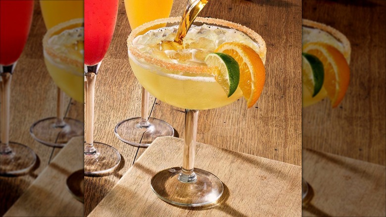 An amber-colored liquid being poured into a margarita glass, garnished with an orange and a lime, on a wooden table.