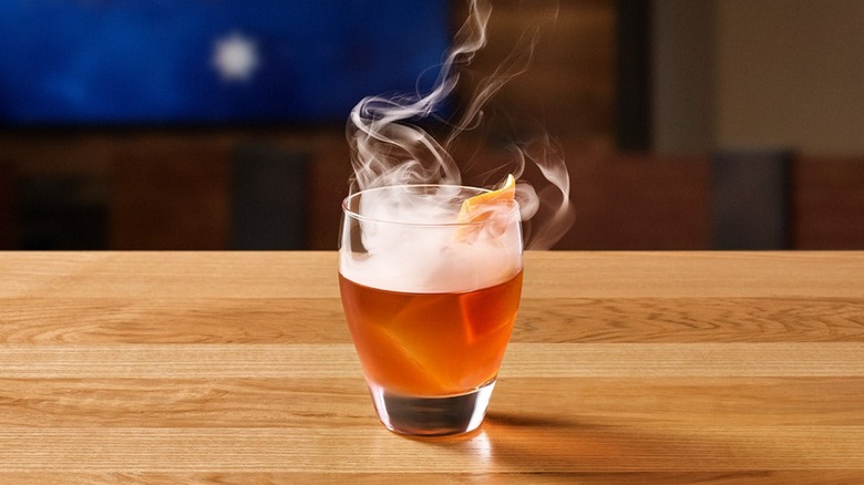 Smoke pours out of an amber-colored cocktail, served in a short glass on a wooden table.