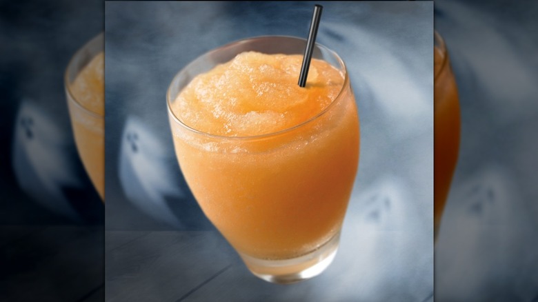 A blended orange cocktail on a wooden table next to a bottle of LaMarca prosecco, a spoon, and a fresh peach.