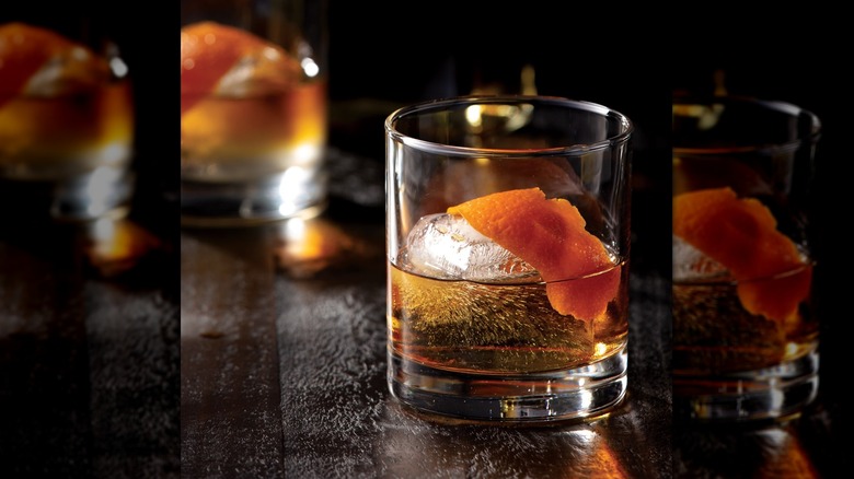 A whiskey cocktail sitting on a wooden bar with an ice cube and an orange slice floating in it. In the background is a white napkin with the text "P.F. Chang's" written on it in black.