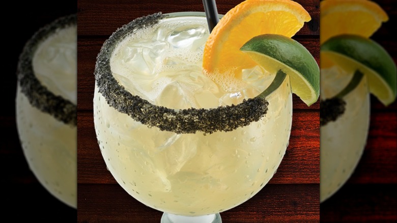 A margarita with a black salt rim, garnished with an orange slice and a lime slice, against a wood grain background.