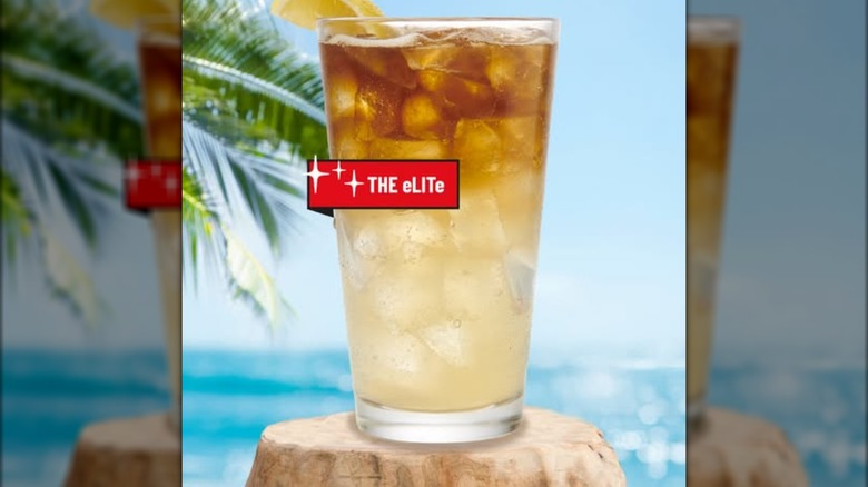 An eLITe sitting on a tree stump with a beachy backdrop. The drink is split down the middle - golden-colored lemonade at the bottom, amber-colored cola at the top.