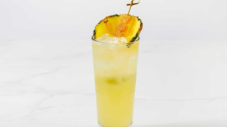 A yellow cocktail in a highball glass, garnished with a pineapple slice.
