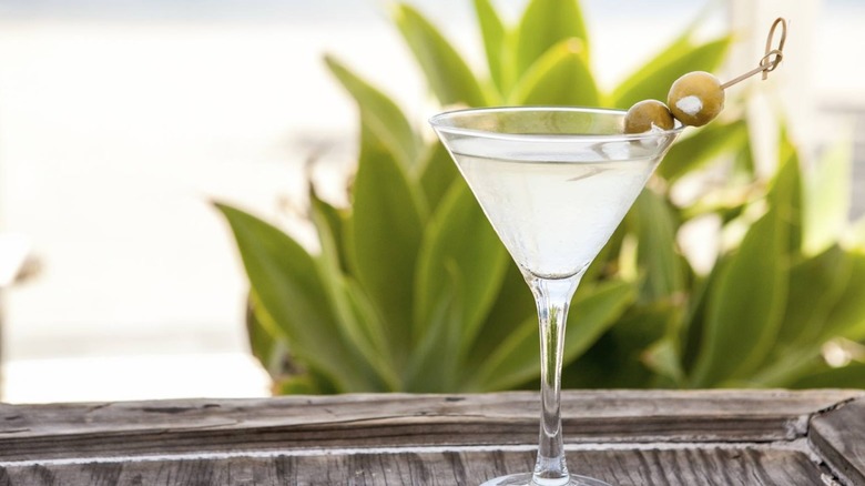 A cloudy martini garnished with two blue cheese-stuffed olives on a wooden table with a green plant in the background.