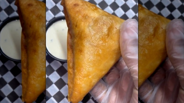 Gloved hand holding sambusa with dipping sauce on checkered paper background