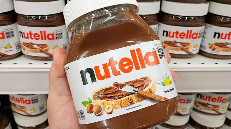 Hand holding a container of Nutella spread with other containers on grocery store shelf in background