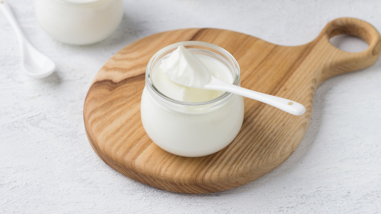 Jar of yogurt on small wooden platter, with spoonful of yogurt on top