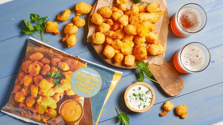 Overhead view of Trader Joe's Breaded Cheddar Cheese Curds bag and baked curds with drinks and sauces on blue table