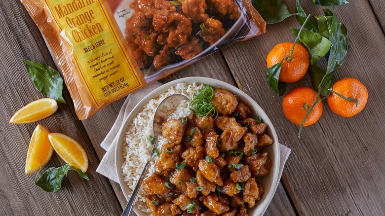 Bag of Trader Joe's Mandarin Orange Chicken on table above a white bowl of orange chicken and rice, mandarins surrounding