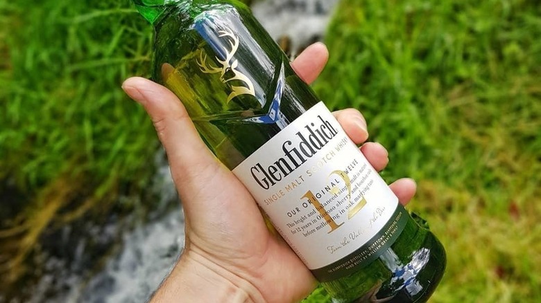 A person holding a bottle of Glenfiddich 12-Year up to the camera with a blurred grassy background