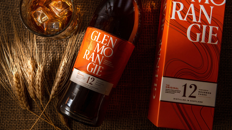 A bottle of Glenmorangie 12-year laid down next to its box, a filled glass, and barley ears