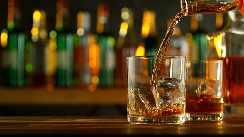 A glass of whisky being poured on a counter with blurred bottles in the background