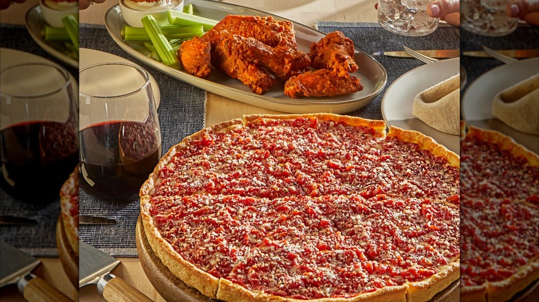 Deep dish pizza on table with glass of wine and platter of chicken wings