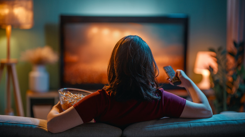 Back view of woman watching TV with remote and popcorn in hands