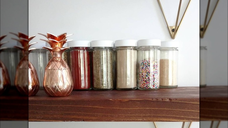 Spice jars filled with spices and sprinkles sit on a wooden shelf.
