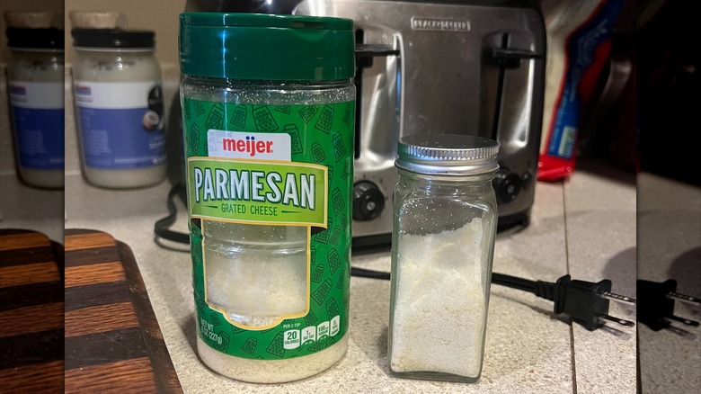 A spice jar holding Parmesan cheese sits on a counter next to a large canister of Parmesan cheese.
