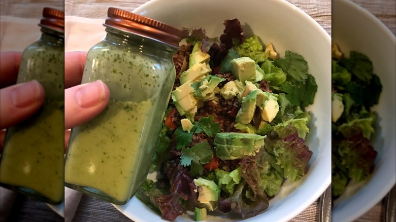 A person holds a spice jar dilled with green salad dressing over a salad of greens and avocado.